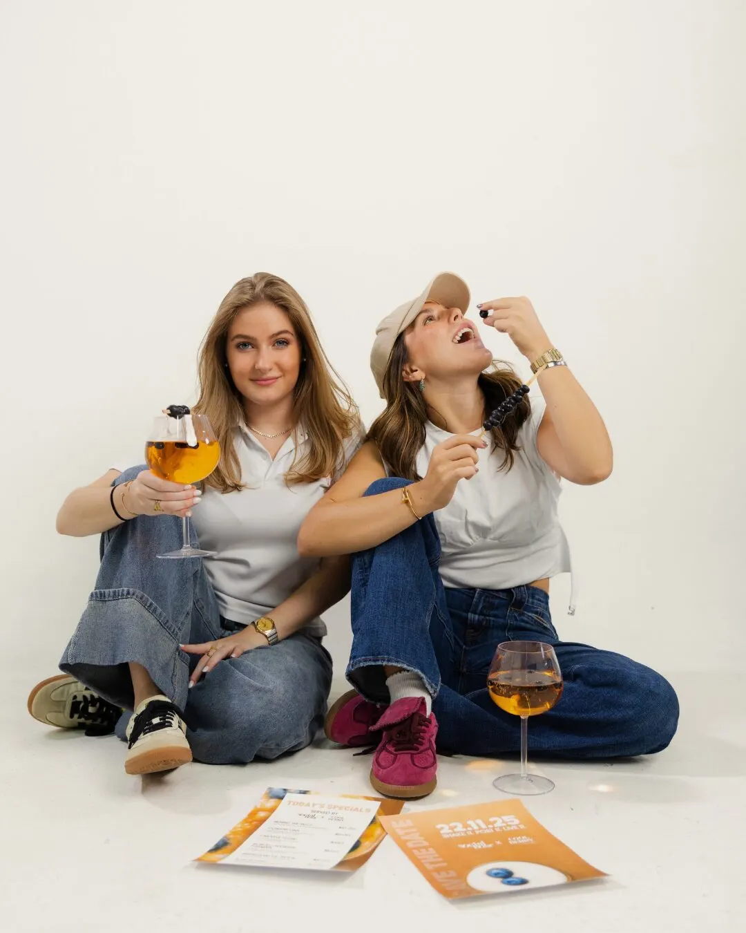 Las dos anfitrionas sentadas en el piso, una con una copa y otra comiendo bayas, con los programas del evento frente a ellas
