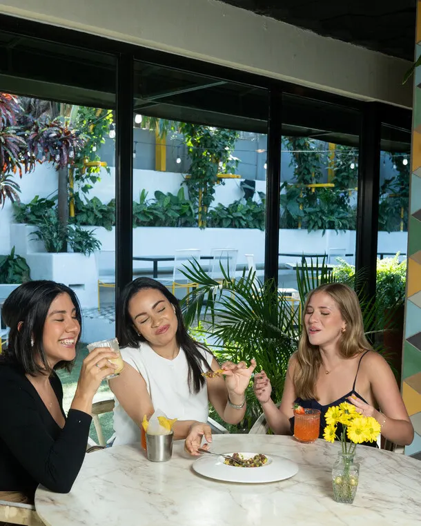 Tres amigas ríen y comparten una mesa de mármol junto a un ventanal con plantas, en Barrio Café.