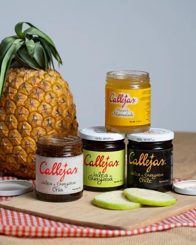 Una piña, tres frascos de jalea Callejas y rodajas de guayaba sobre una tabla de madera.
