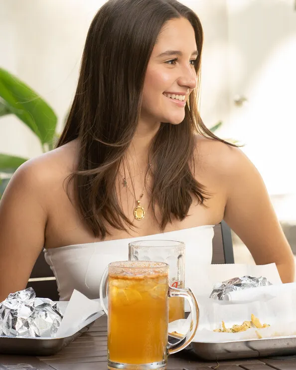 Joven sonriente junto a una jarra de bebida y papel de aluminio de hamburguesa, en una terraza de TBG.