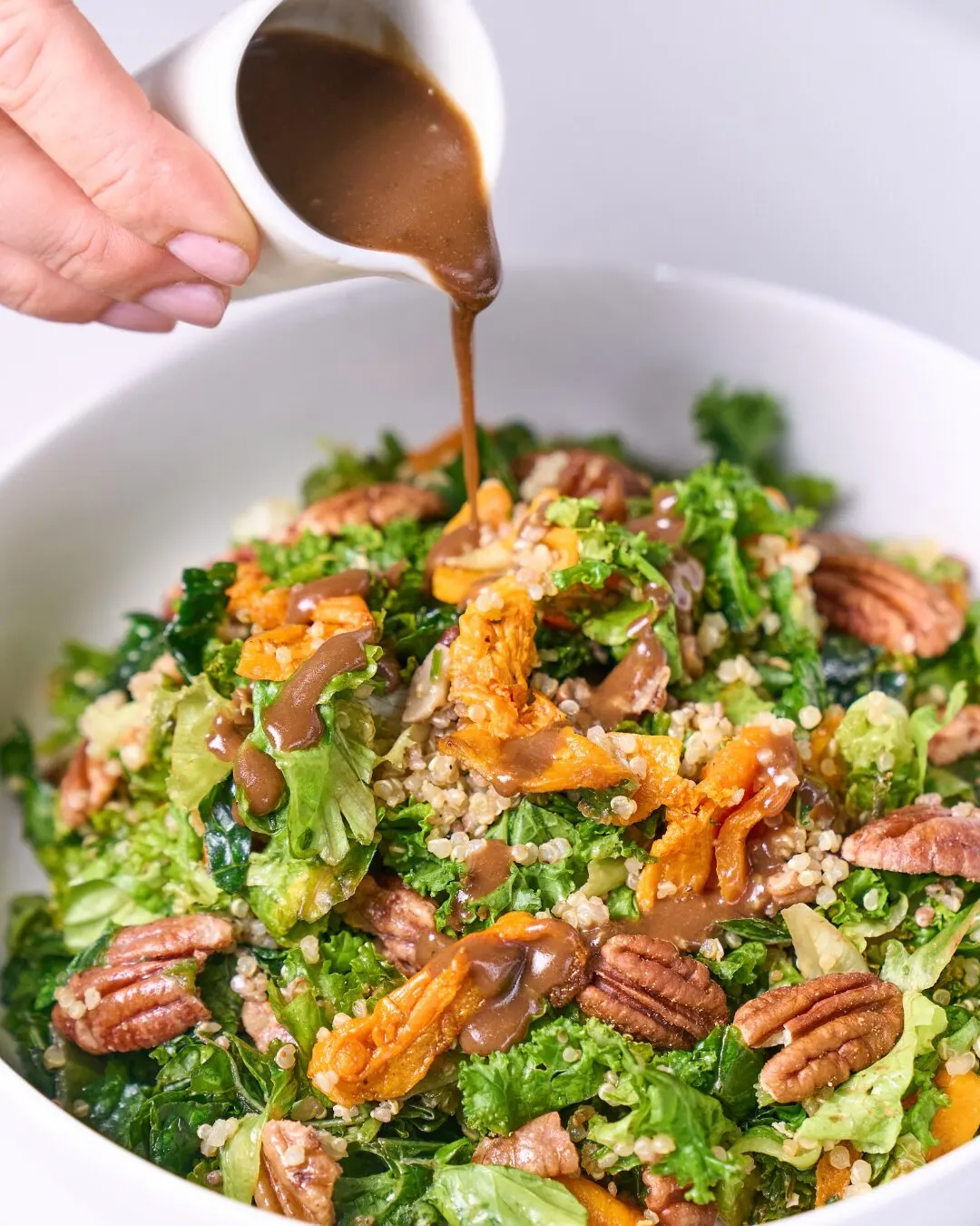 A hand pouring a dark dressing over a kale salad with pecans and carrot in a white bowl.