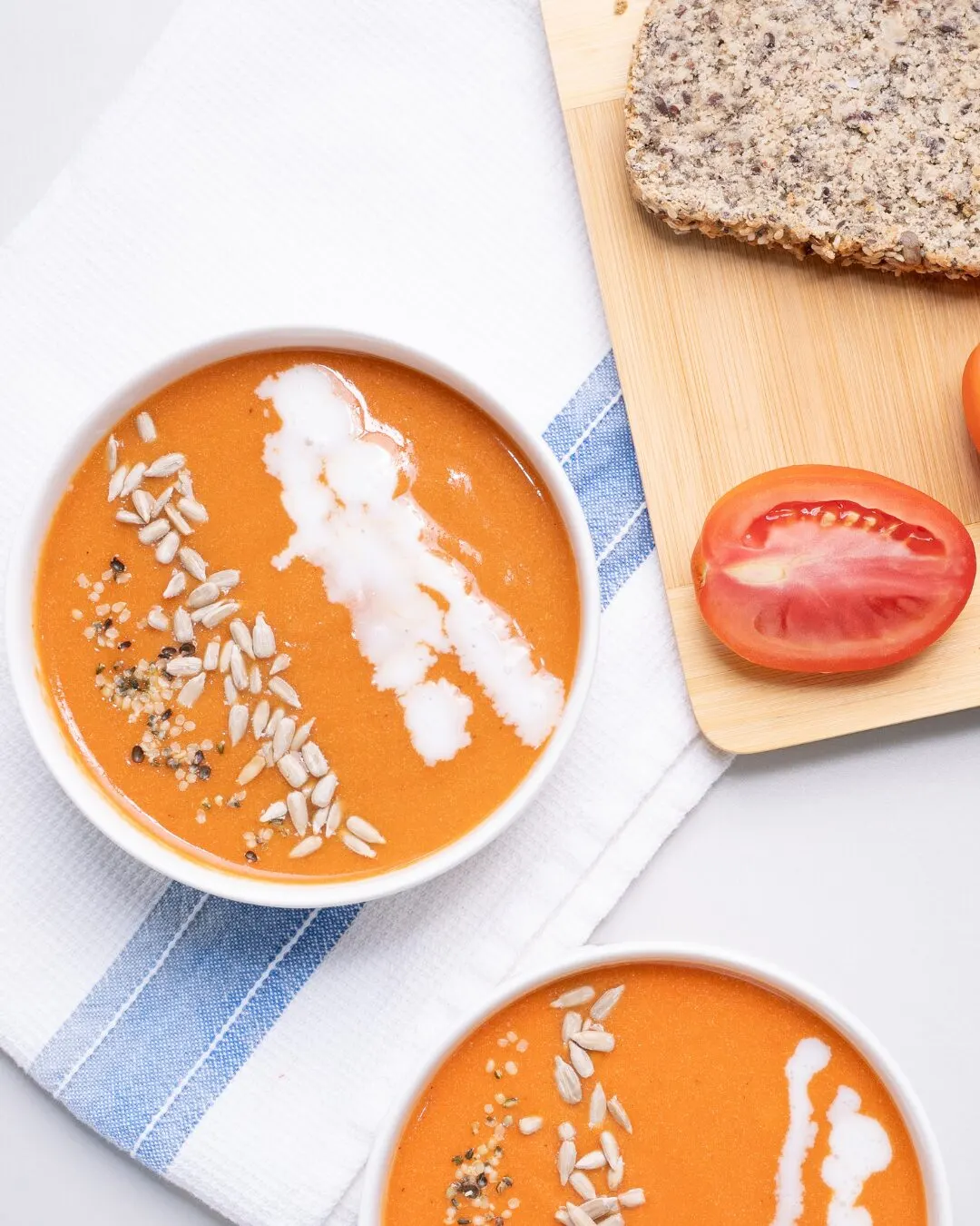 Two bowls of tomato soup with seeds and cream, with a tomato and bread slices beside them.