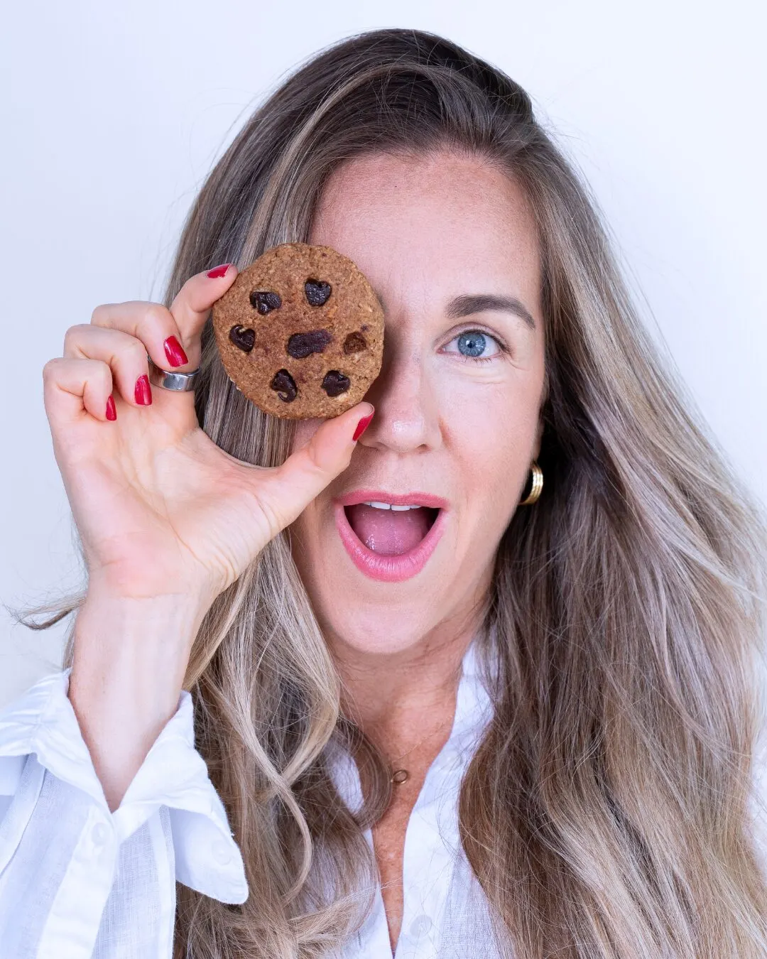 A woman with long hair holding a chocolate cookie in front of one eye, mouth open in surprise.