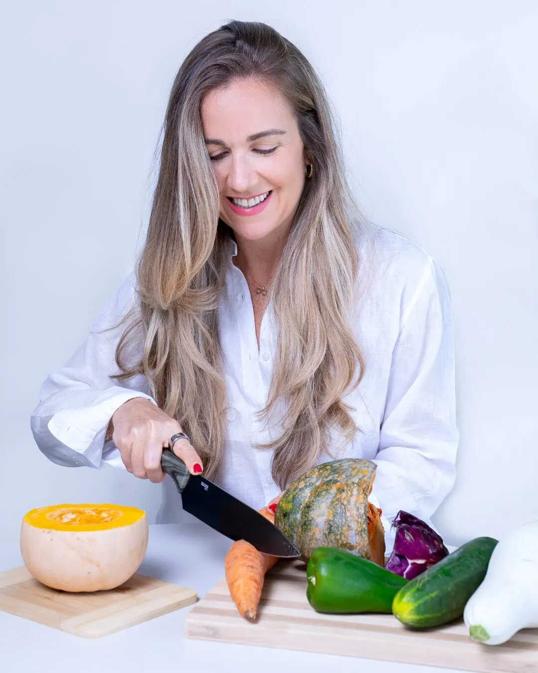 A woman in a white blouse cutting squash and vegetables on a wooden board.