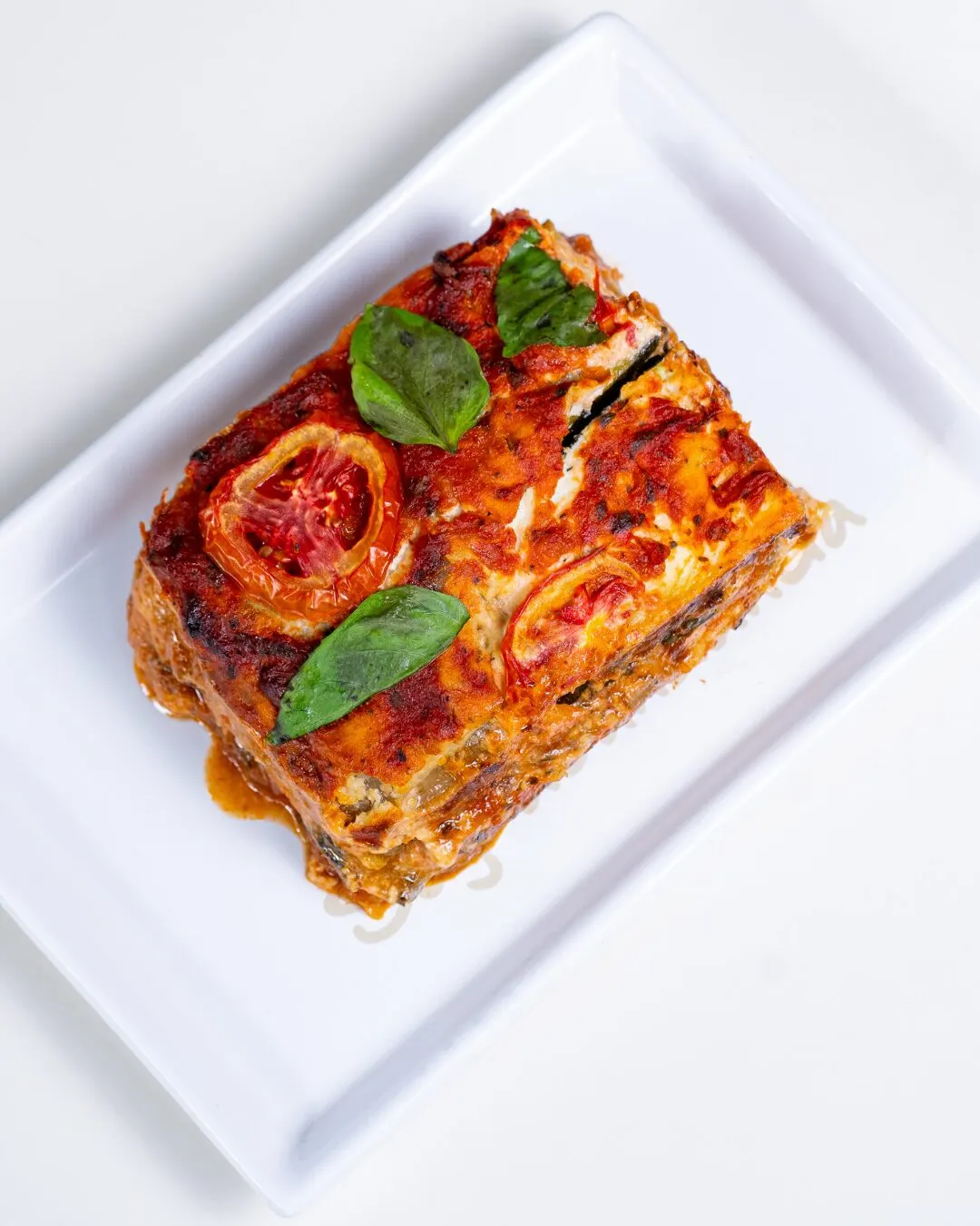 Vegetable lasagna with basil and tomato on a square white plate.
