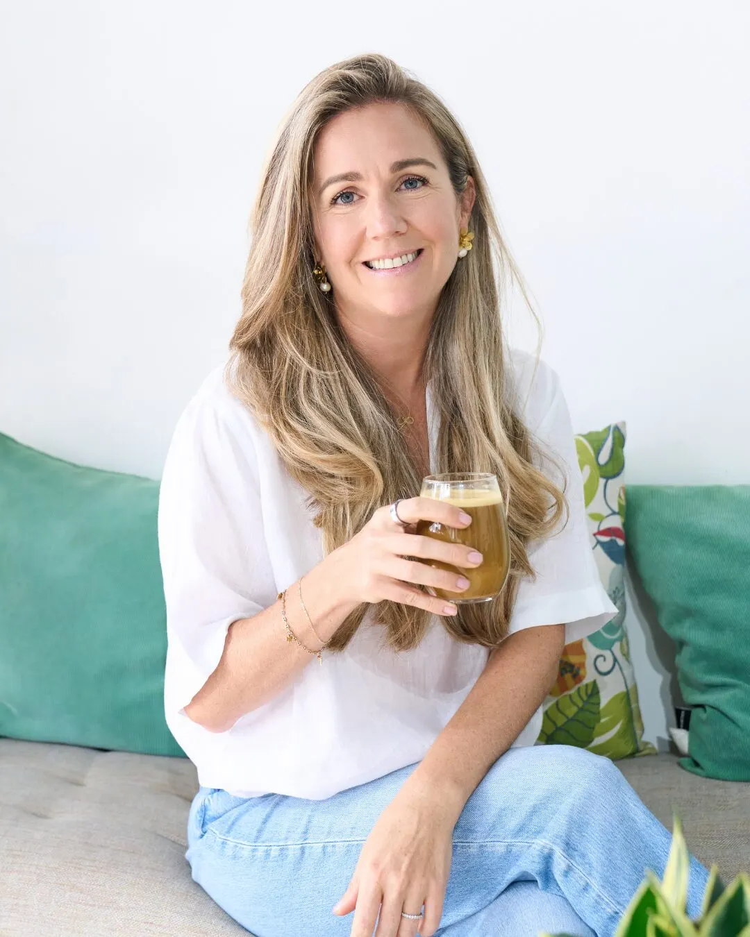 A woman with long blonde hair smiling on a green sofa, holding a smoothie glass.