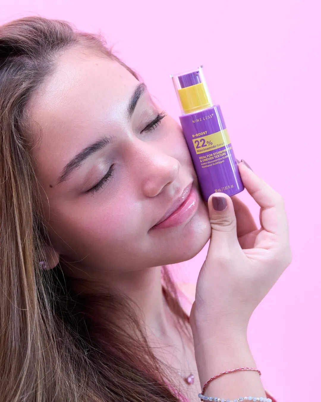 A young woman with closed eyes holding a purple sunscreen stick against her cheek, on a pink background.