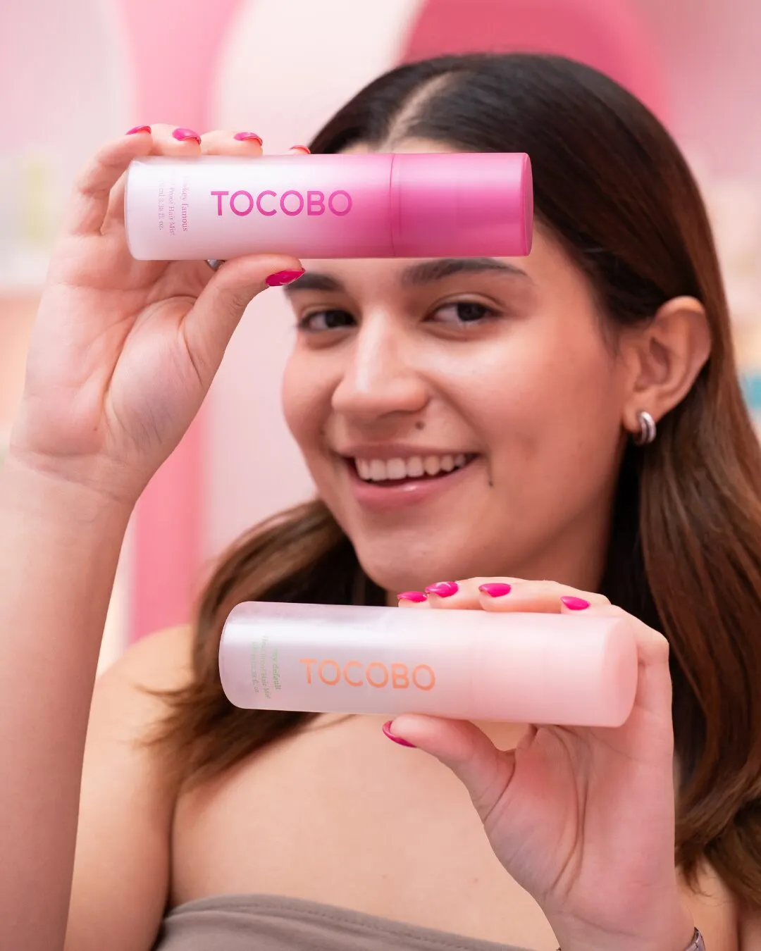 A smiling woman holding two pink tubes, one above her head and one at chin height.