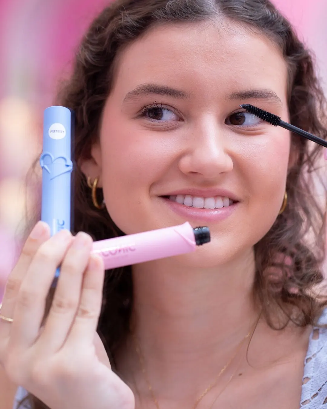 A smiling woman applying mascara, with a blue container in her other hand.