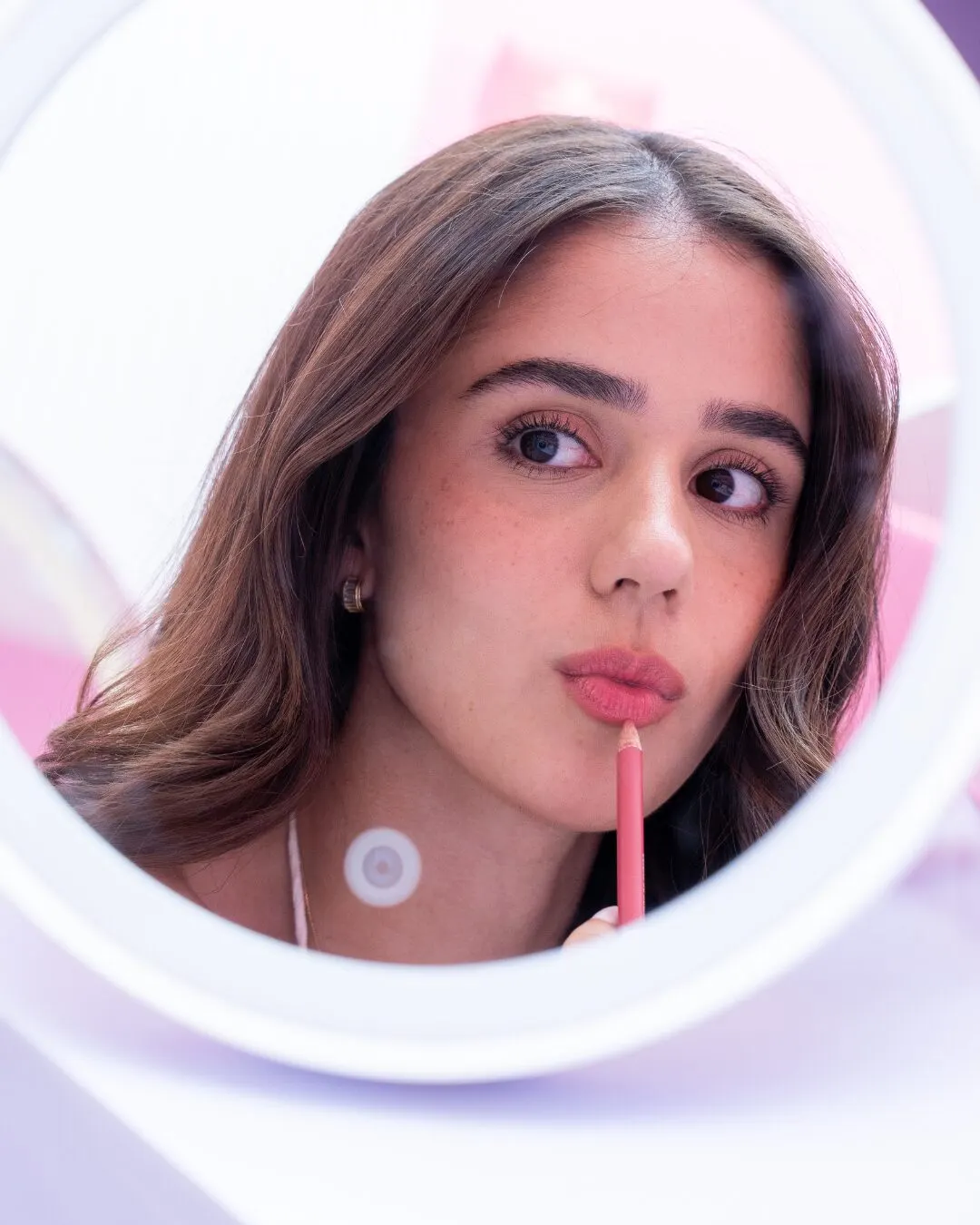 A young woman at a round lit mirror, applying a pink lip product.