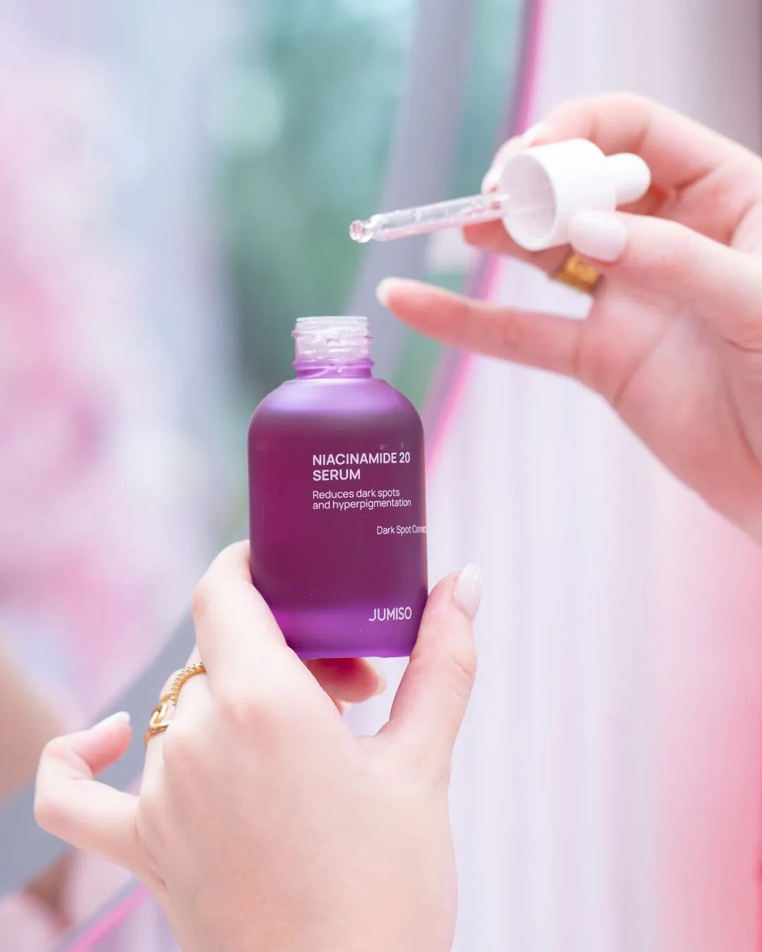 Hands holding a dropper over a purple serum bottle, against a pink background.