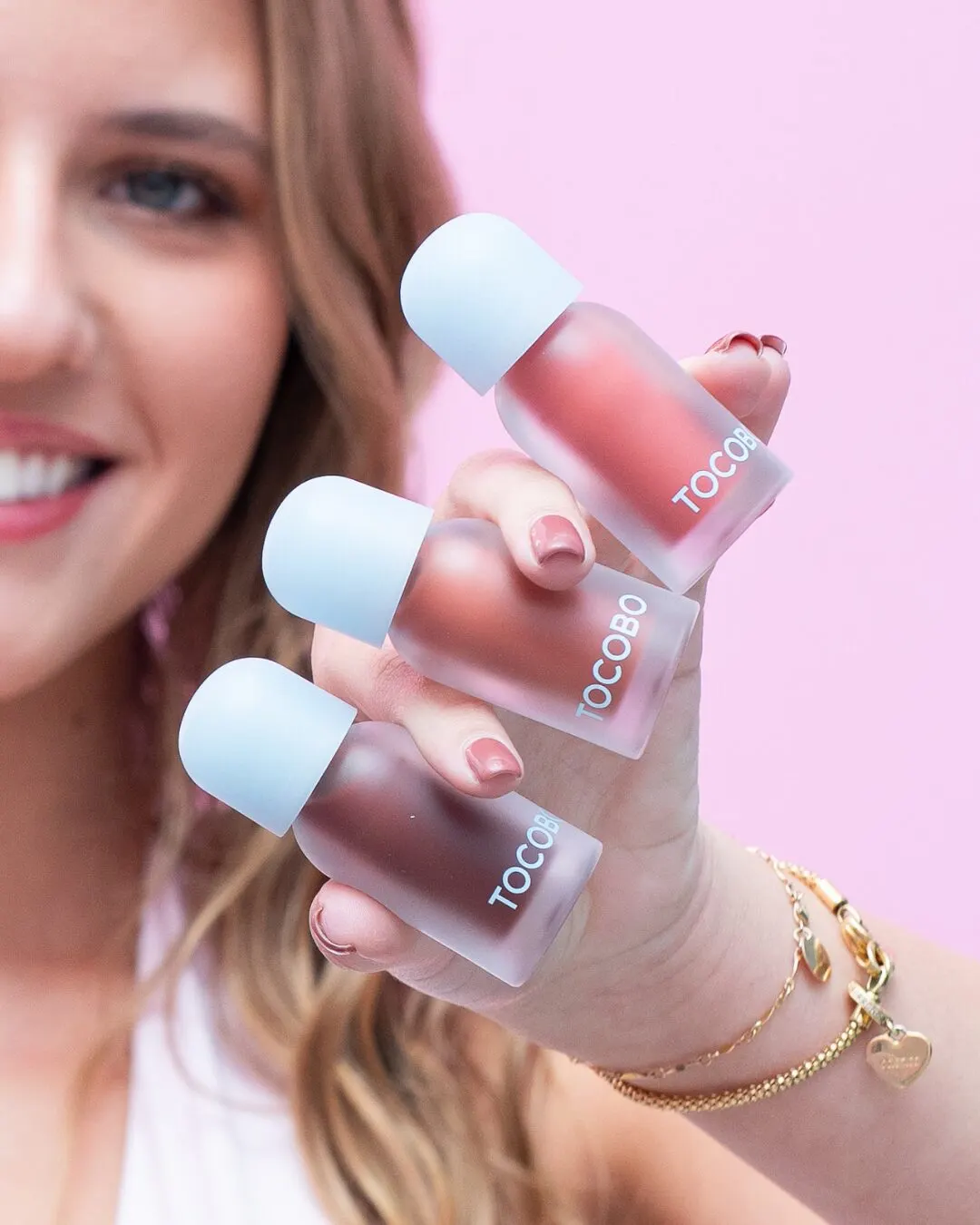 A hand holding three lip tints with light-blue caps in front of a smile.