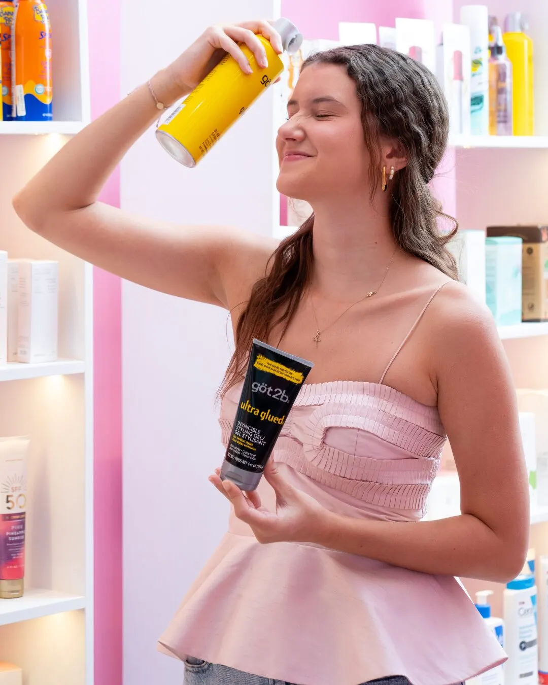 A young woman in a pink top holding a yellow spray can up high and a black tube in her other hand, beside a product shelf.