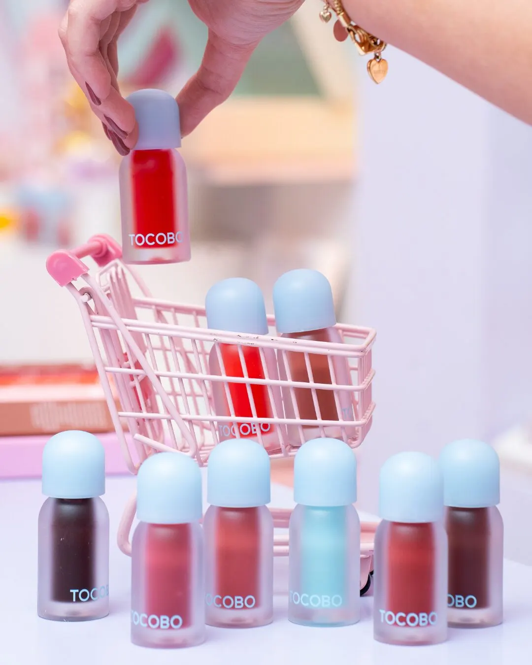 A miniature pink shopping cart surrounded by lip tints in several colors, with a hand holding one.
