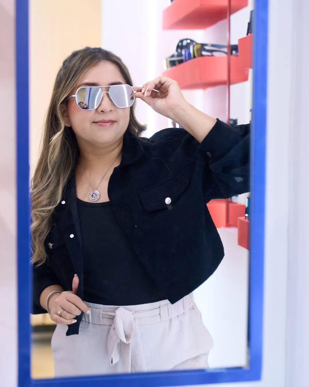 A woman trying on silver sunglasses in front of a blue-framed mirror.