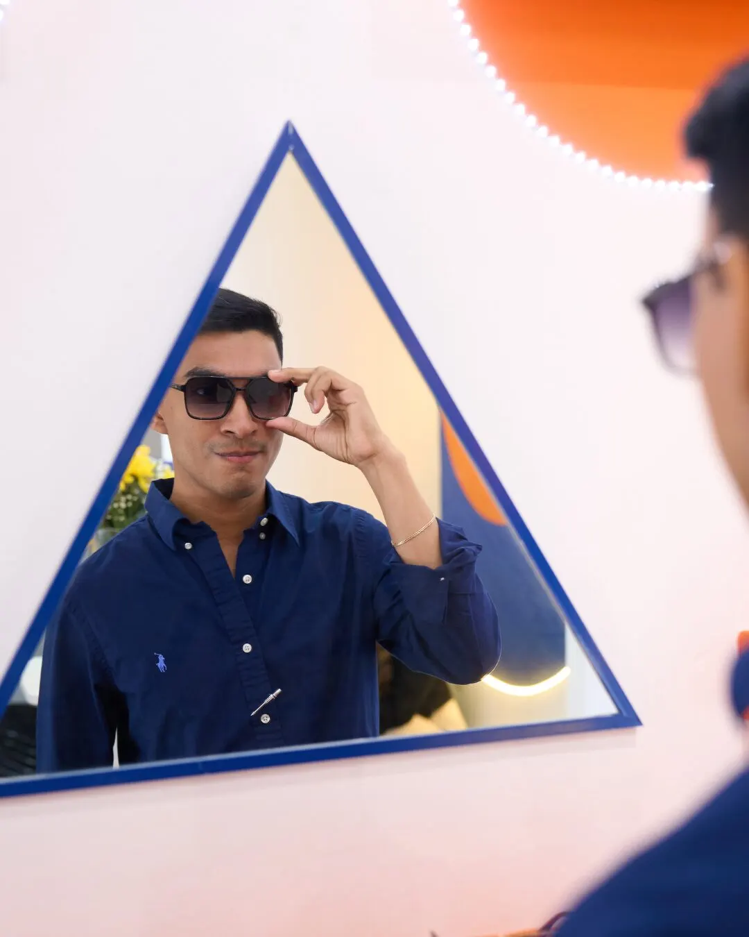 A man in dark sunglasses reflected in a blue-framed triangular mirror.