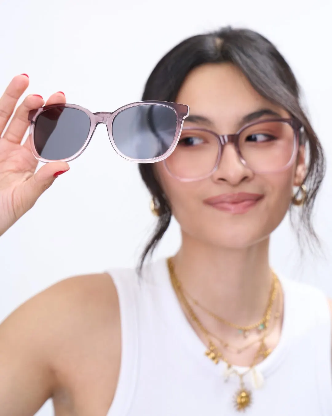 A young woman in a white top wearing pink-framed glasses and holding gray-lens sunglasses above her forehead.