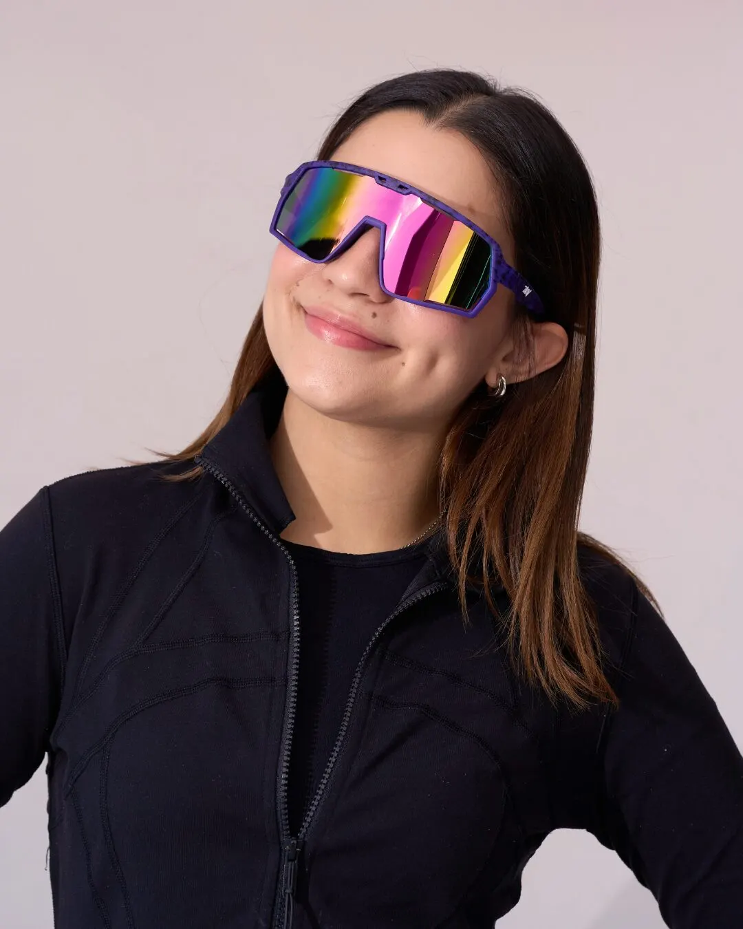A young woman in sporty sunglasses with iridescent lenses and a black jacket, smiling.