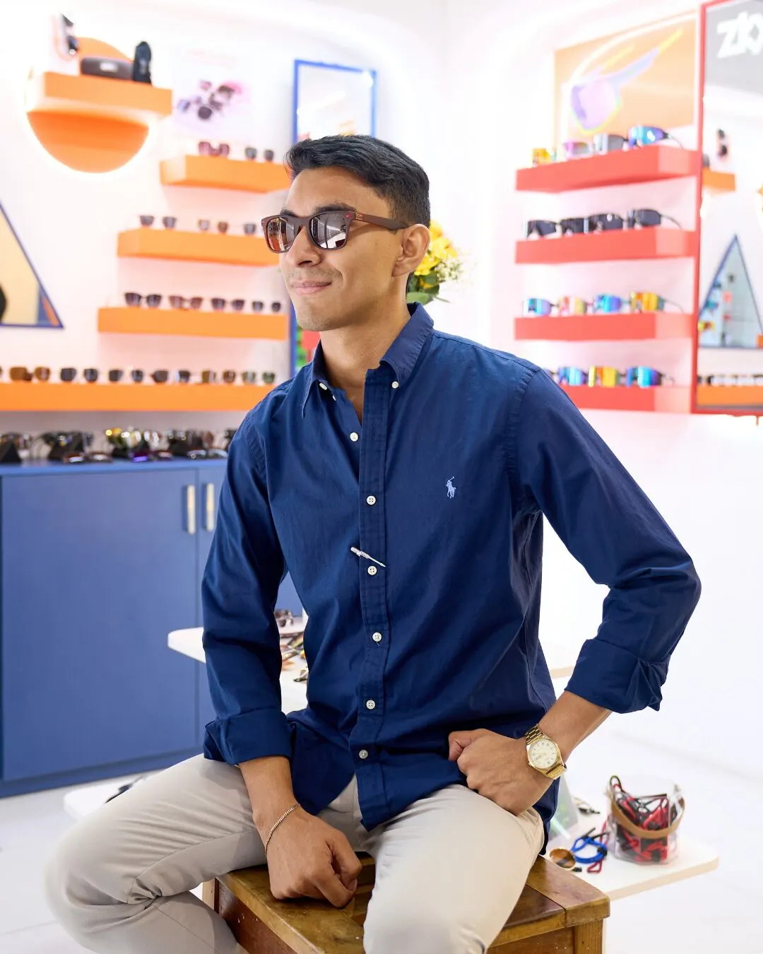 A man in a navy shirt and dark sunglasses sitting on a stool in the store, with orange shelves full of sunglasses behind him.