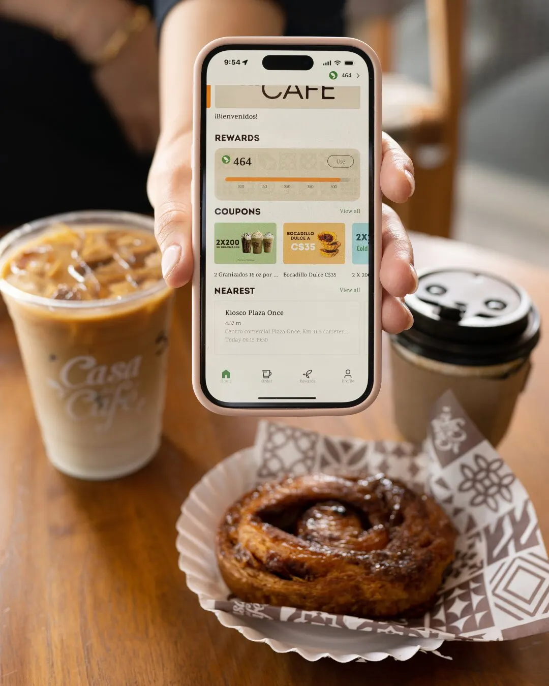 Mano mostrando la app de Casa del Café junto a un café helado y un pan de canela.