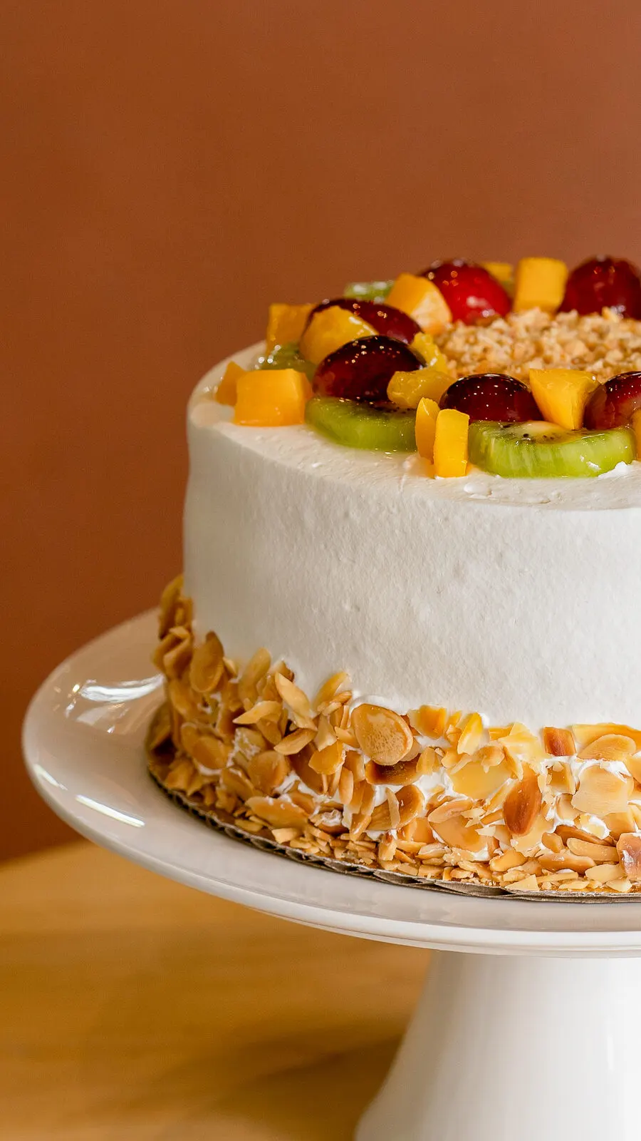 Pastel blanco con frutas y almendras laminadas sobre un pedestal, frente a una pared color terracota.