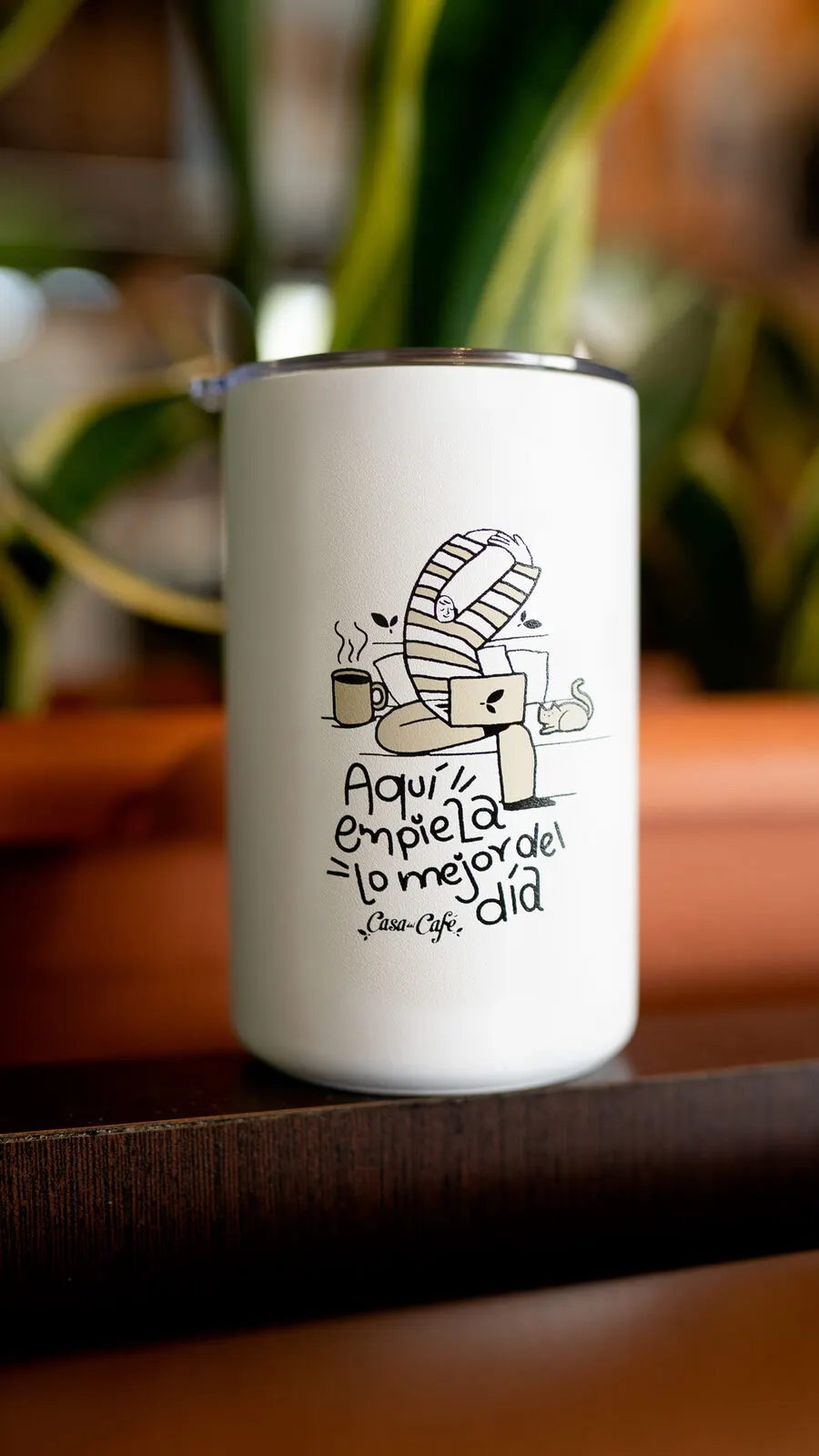 Termo blanco ilustrado de Casa del Café sobre una mesa, con una planta detrás.