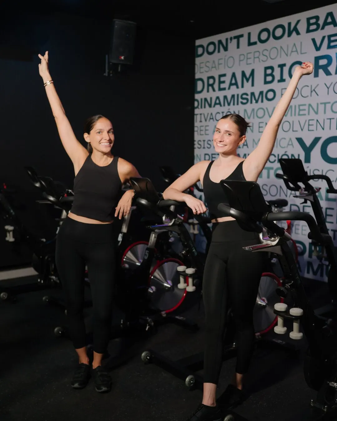 Dos instructoras celebrando con los brazos arriba entre las bicicletas del estudio.