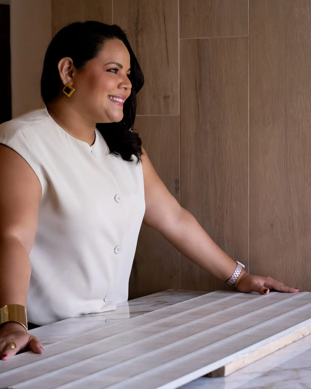 Mujer sonriente con la mano apoyada sobre un panel de mármol, frente a una pared de madera.