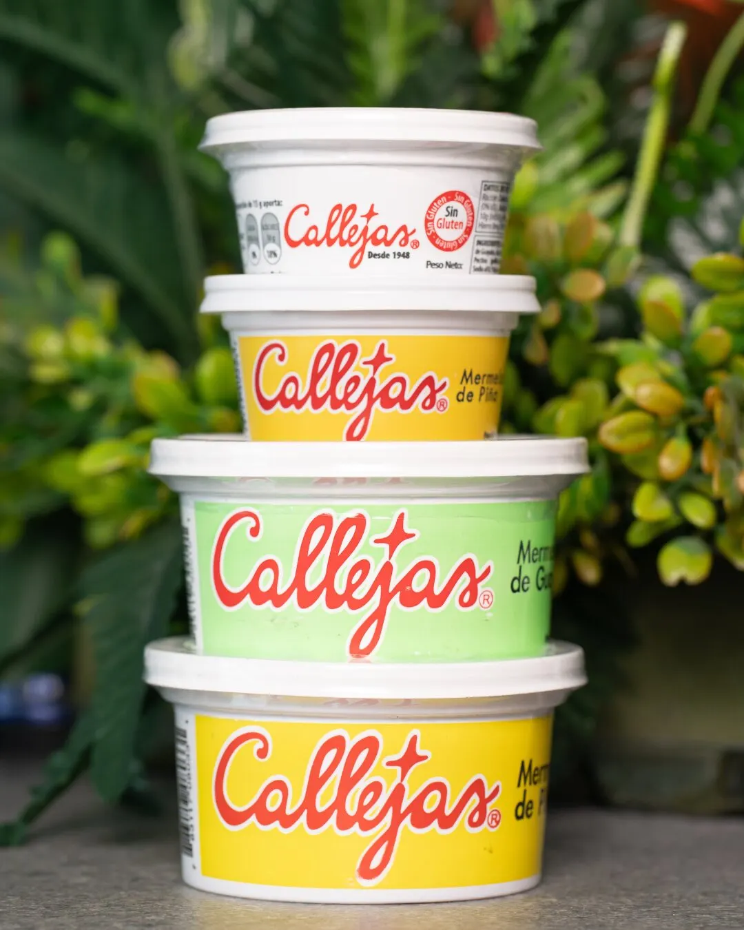 Potes de jalea Callejas apilados en colores amarillo, verde y blanco, entre plantas.