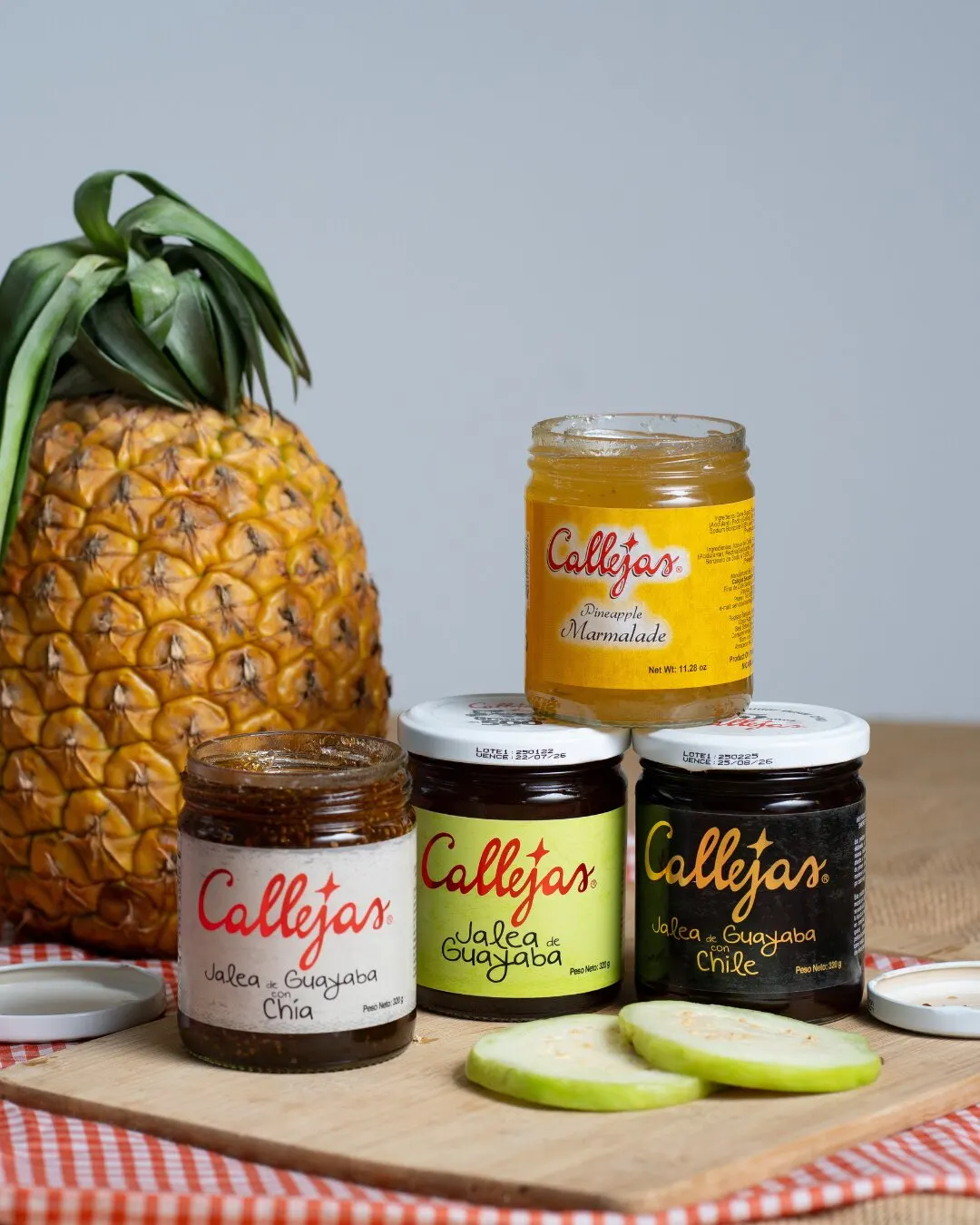 Una piña, frascos de jalea Callejas y rodajas de guayaba sobre un mantel de cuadros.