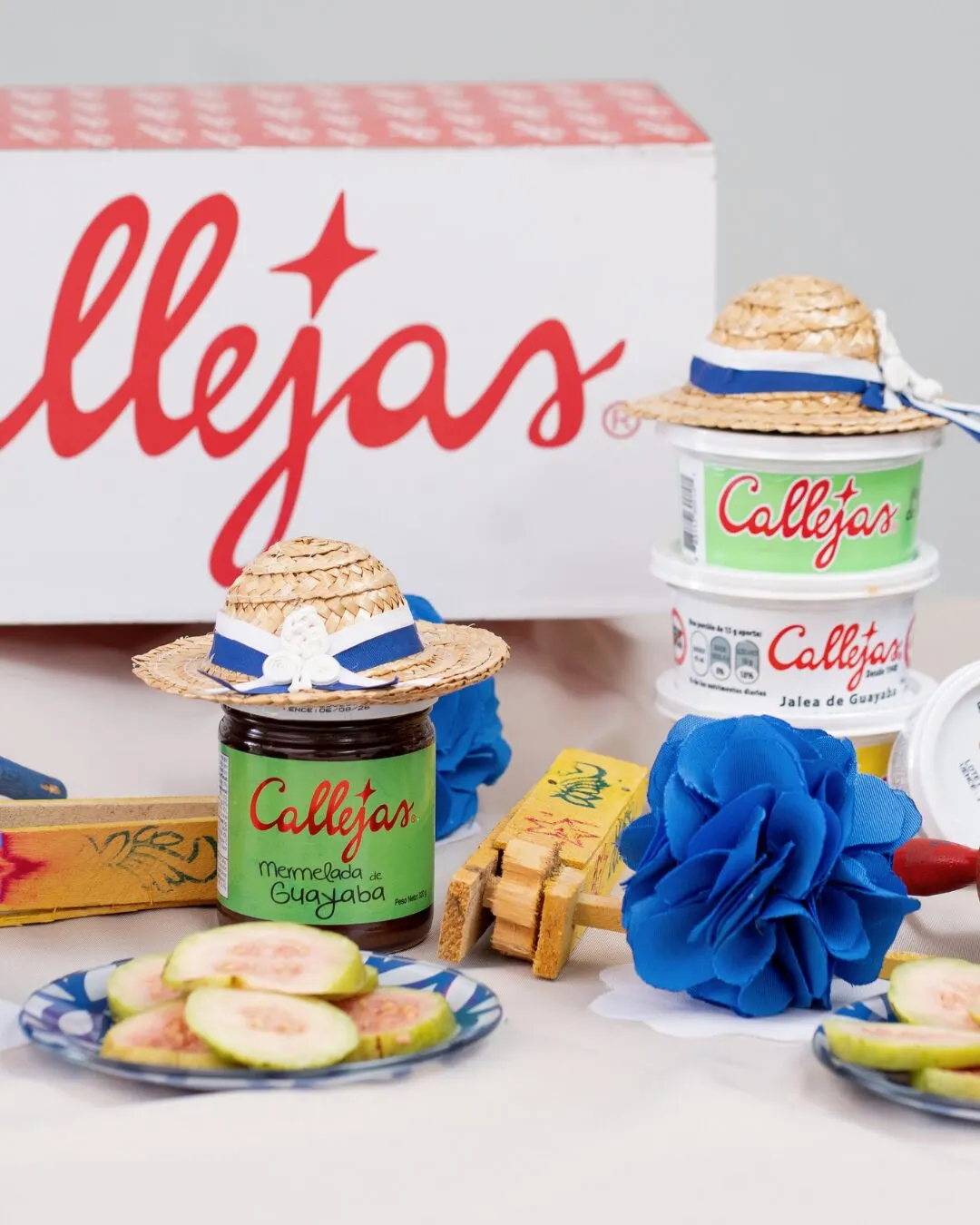 Caja con el logo de Callejas, frascos y vasitos de jalea, un sombrerito de paja y flores azules sobre una mesa con rodajas de guayaba.