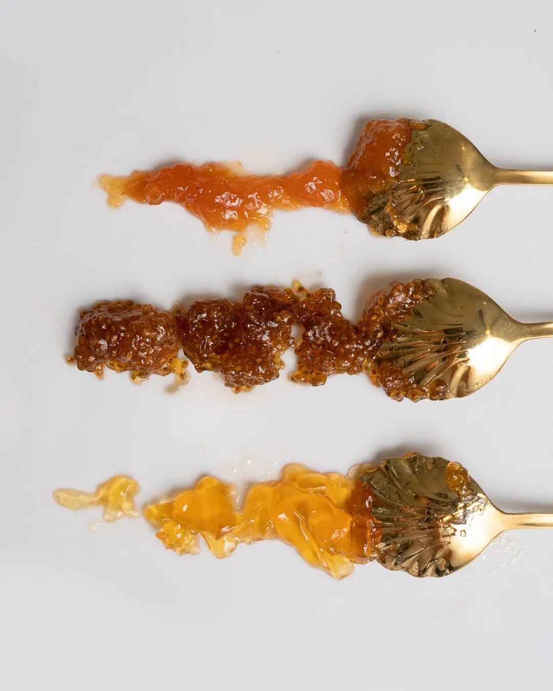 Tres cucharas doradas con untados de mermelada, jalea y miel sobre un fondo gris claro.