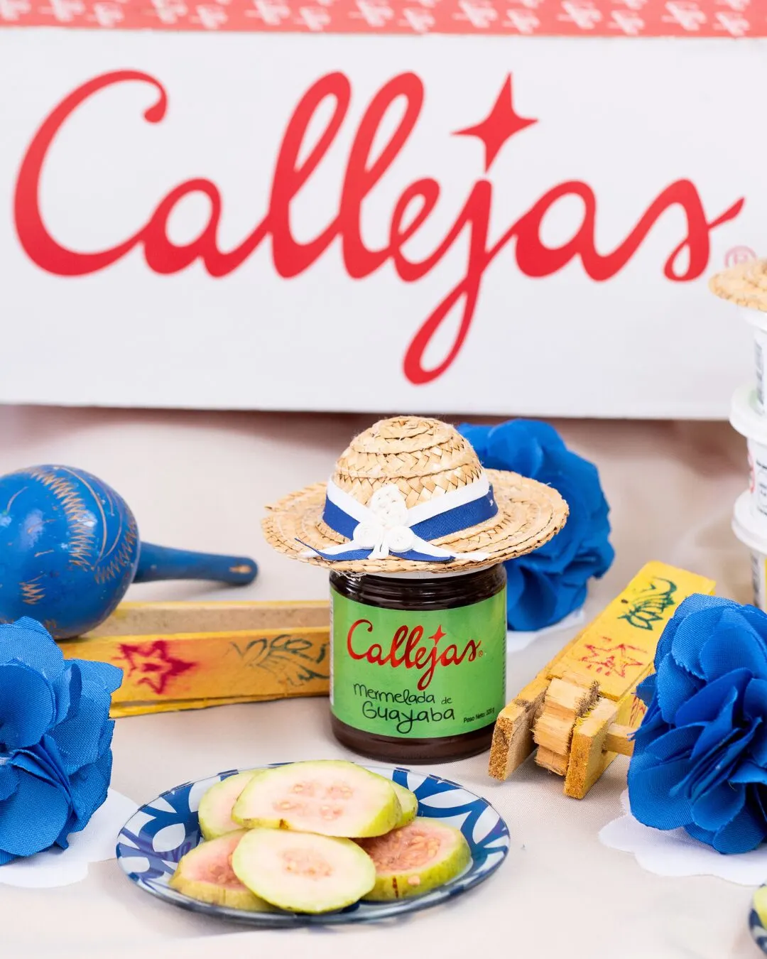 Primer plano de la caja con el logo de Callejas, maracas azules, un frasco de mermelada, un sombrerito de paja y un plato de guayaba.