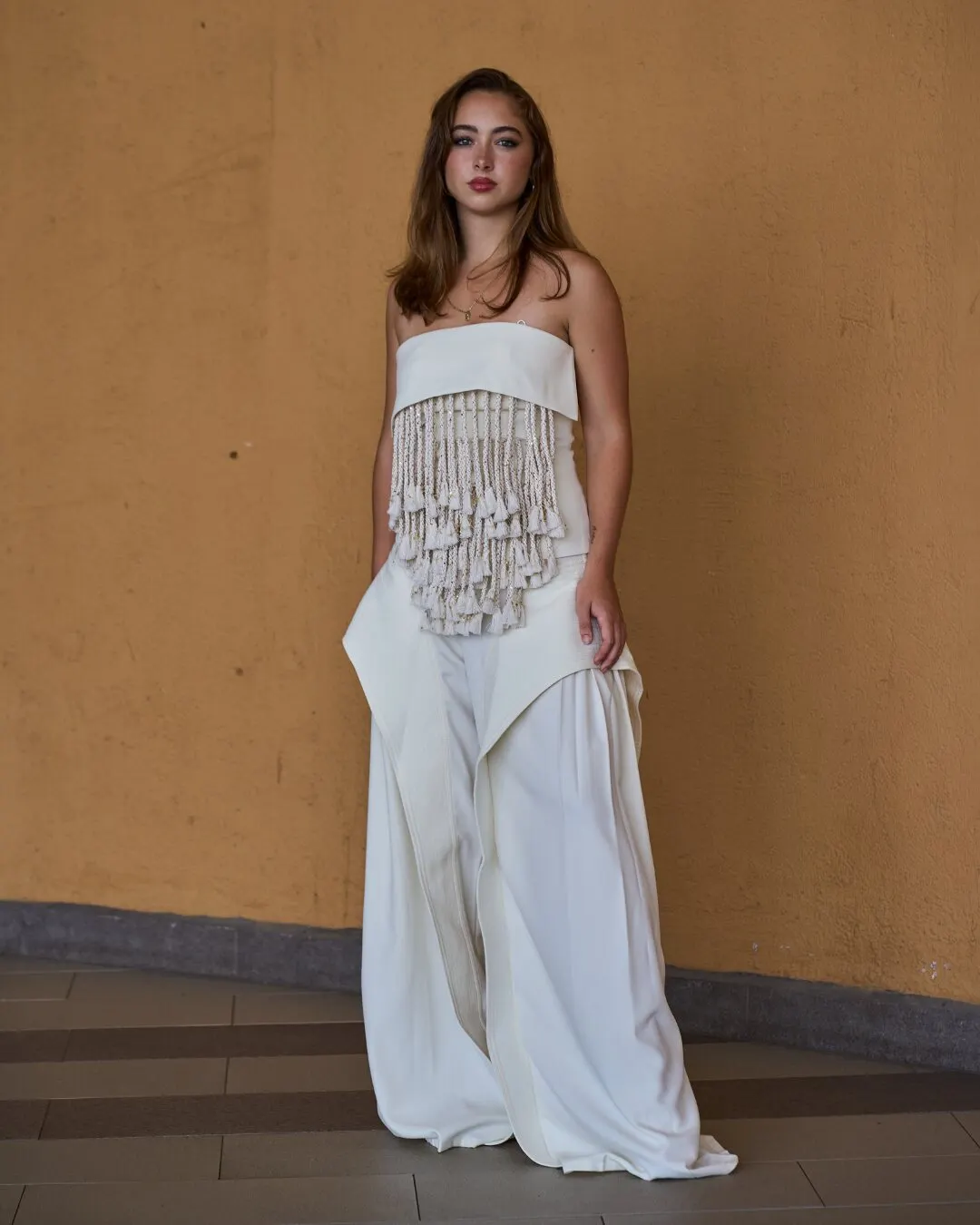 Modelo con top de flecos y pantalón ancho color crema, frente a una pared ocre.