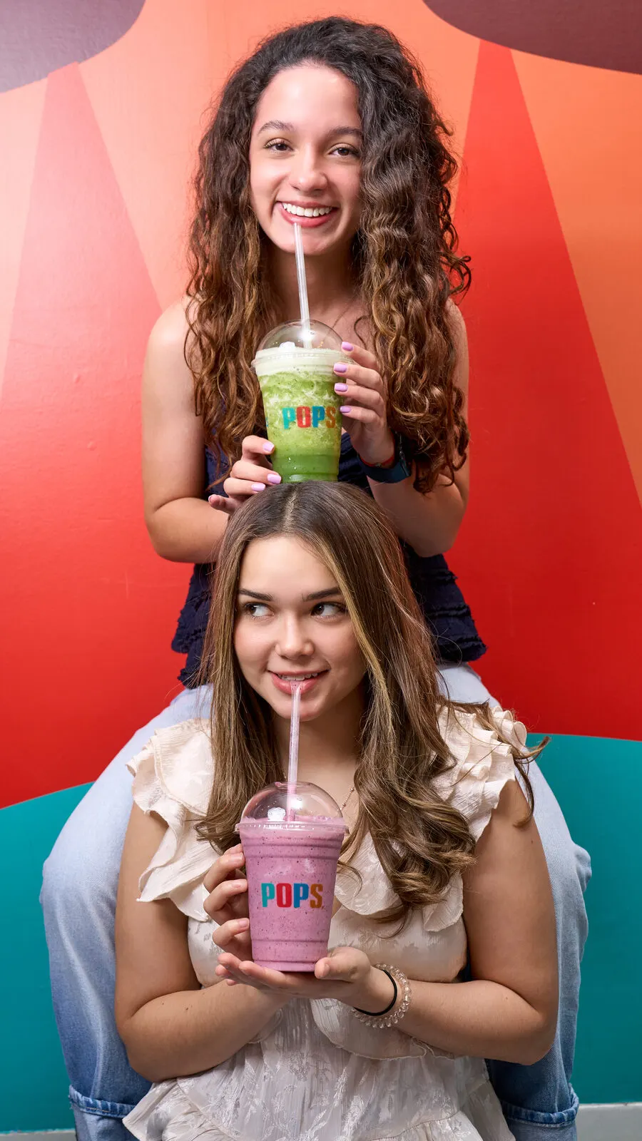 Dos chicas sonrientes con malteadas POPS frente a un mural de colores, una detrás de la otra.