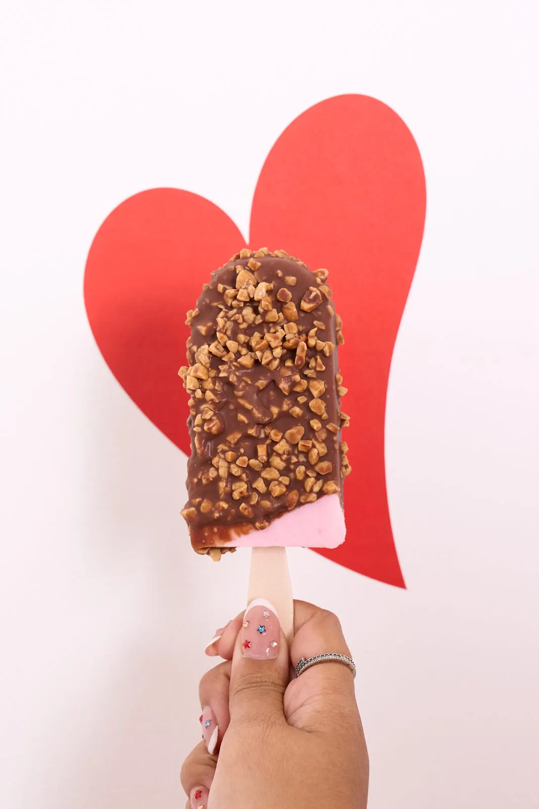 Paleta de chocolate con trocitos frente a un corazón rojo de papel, sostenida por una mano.