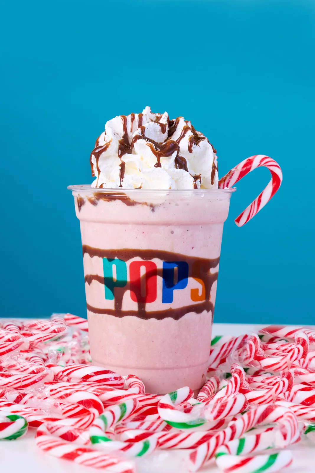 Malteada rosada con crema batida, salsa de chocolate y un bastón de dulce, sobre un fondo turquesa.