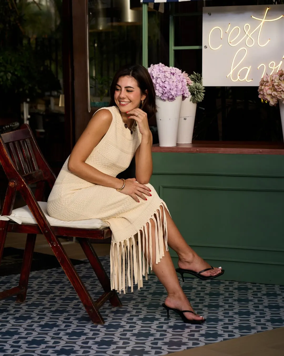 Modelo con un vestido tejido crema de flecos, sentada en una silla de madera junto a hortensias y un letrero de neón.
