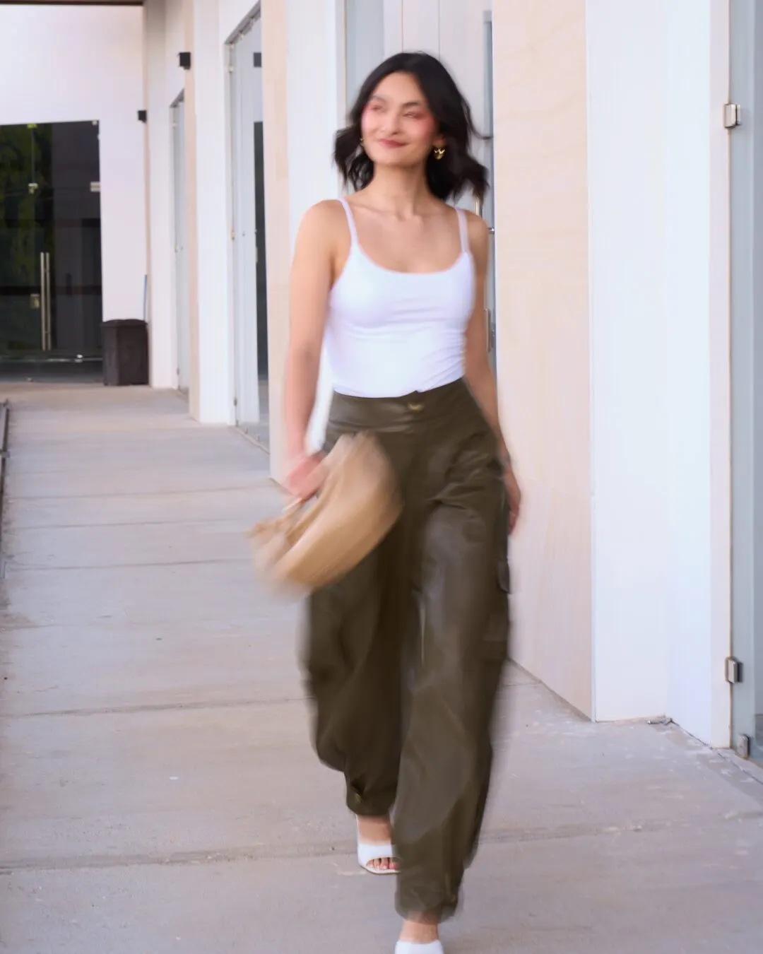 Modelo caminando con un top blanco y pantalón verde oliva, con un bolso en la mano, por un pasillo exterior.