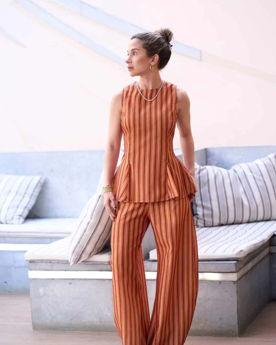 Modelo de perfil con un conjunto de rayas naranja sin mangas, junto a un sofá con cojines de rayas.