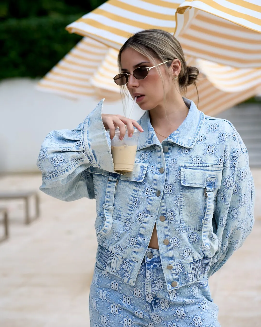 Modelo con lentes de sol y chaqueta de mezclilla clara estampada, con un café helado en la mano, bajo unas sombrillas de rayas.