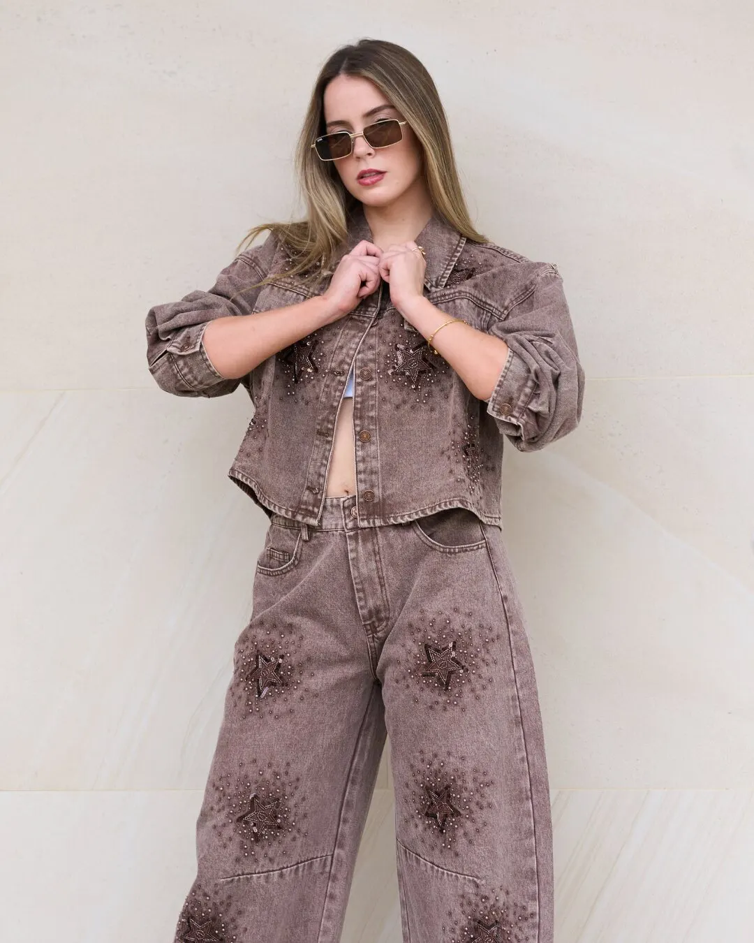 Modelo con gafas y un conjunto de chaqueta y pantalón de mezclilla café con bordados de estrellas, frente a una pared crema.