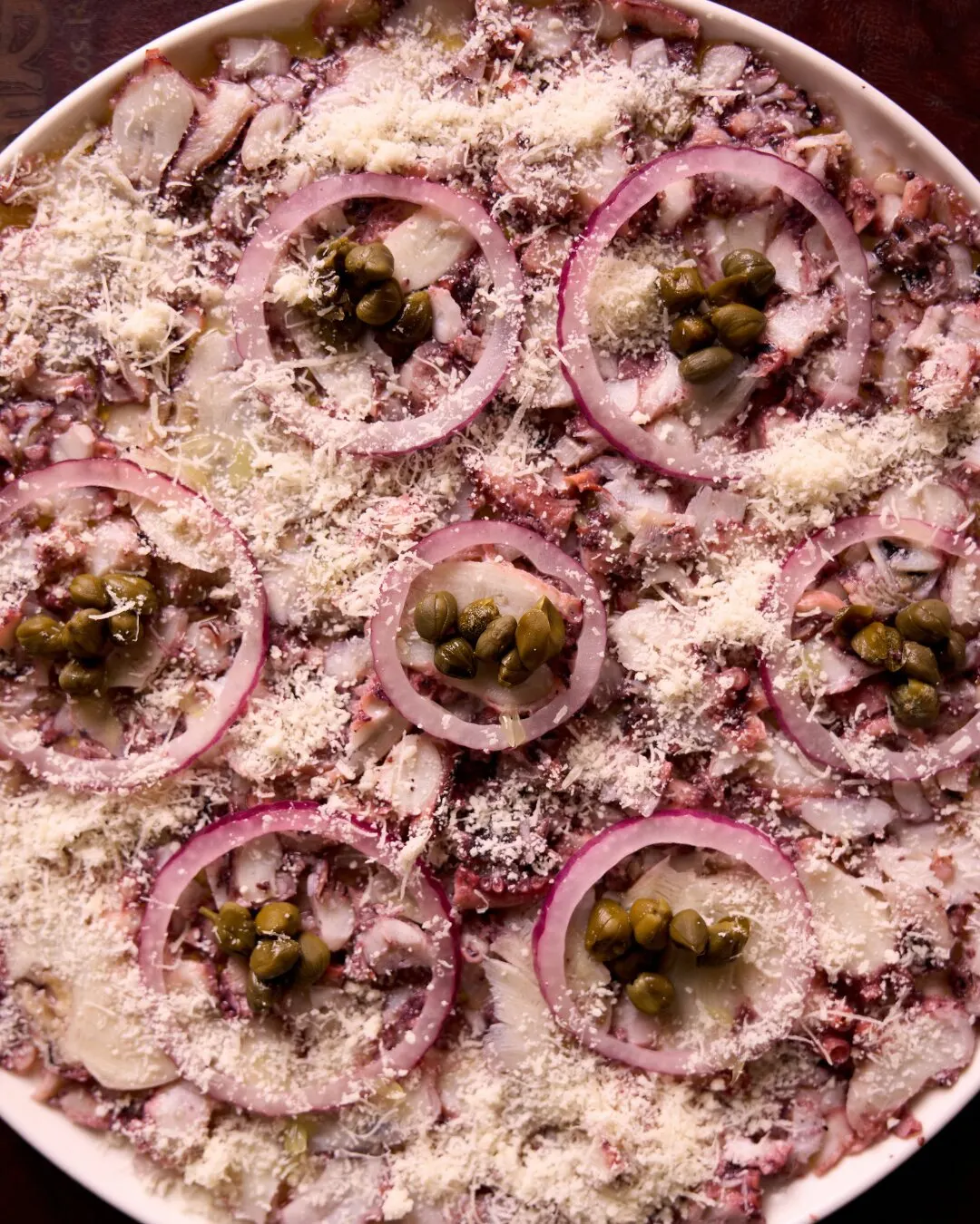 Plato redondo visto desde arriba, con láminas finas cubiertas de aros de cebolla morada, alcaparras y queso rallado.