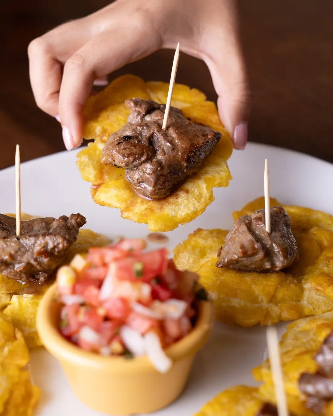 Mano levantando un tostón con carne en un palillo, sobre un plato con pico de gallo.