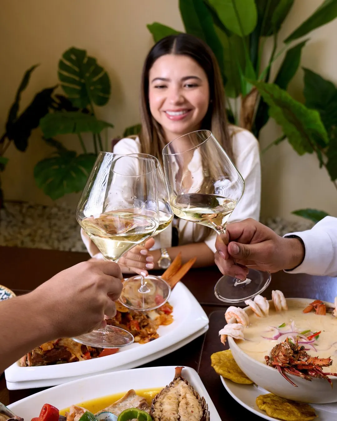 Brindis con copas de vino blanco sobre una mesa con platos de mariscos, con una mujer sonriendo al fondo.