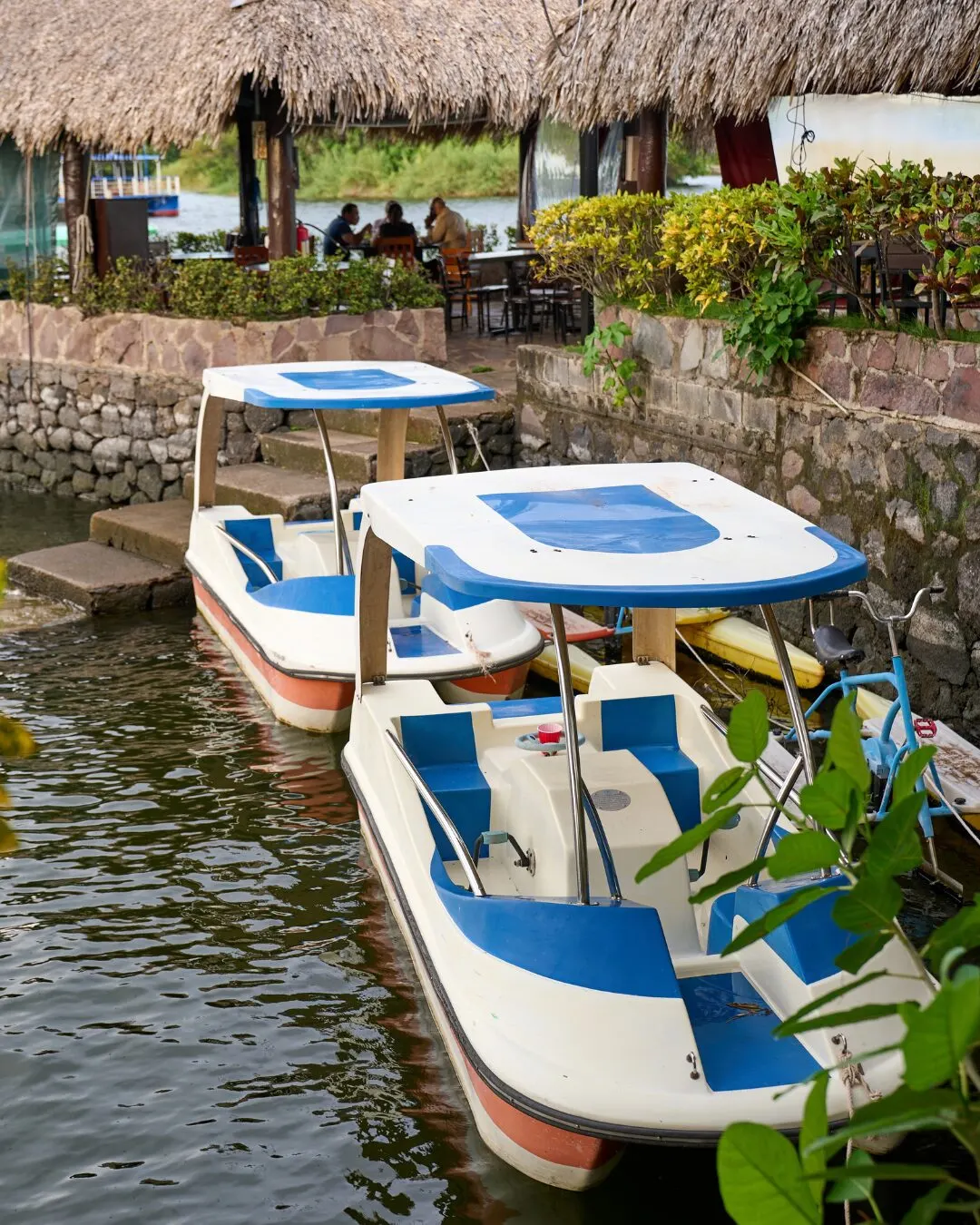Two white-and-blue pedal boats moored on the water in front of a thatched-roof dock.