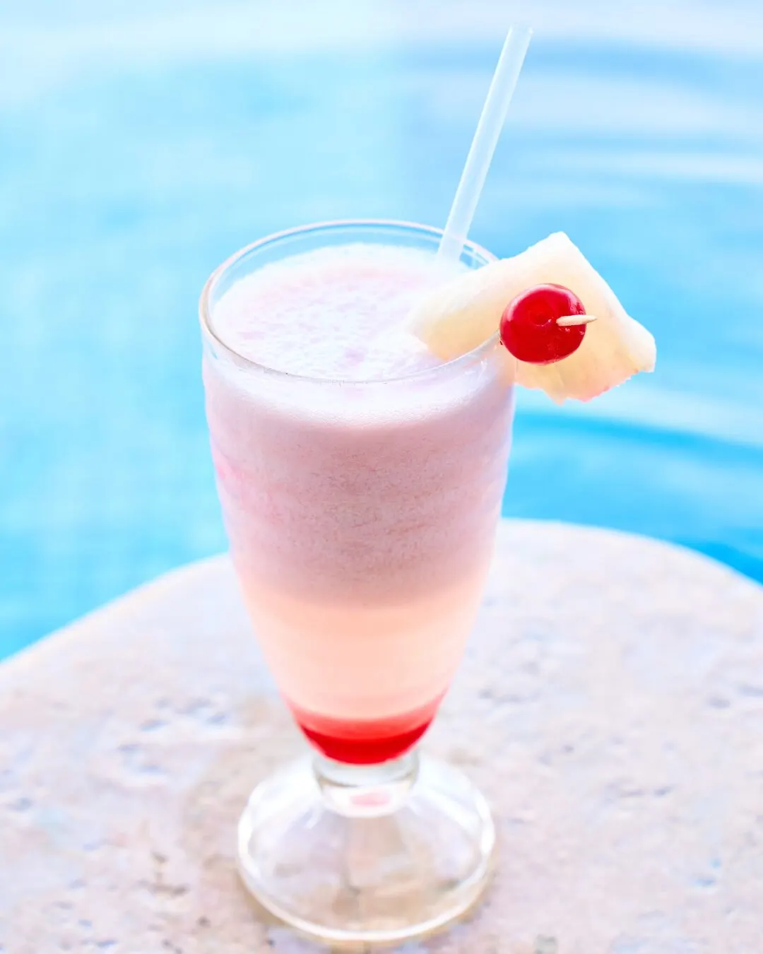 A pink shake topped with a cherry and pineapple, on the edge of a pool.