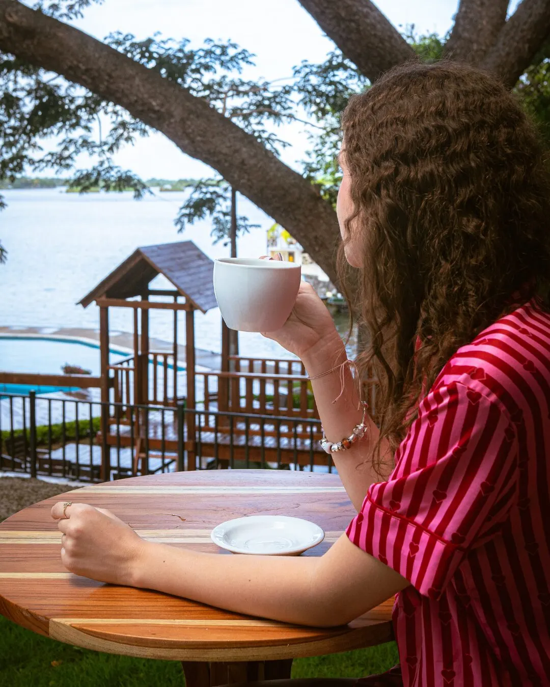 A young woman in a red striped shirt drinking coffee at a wooden table by the lake.