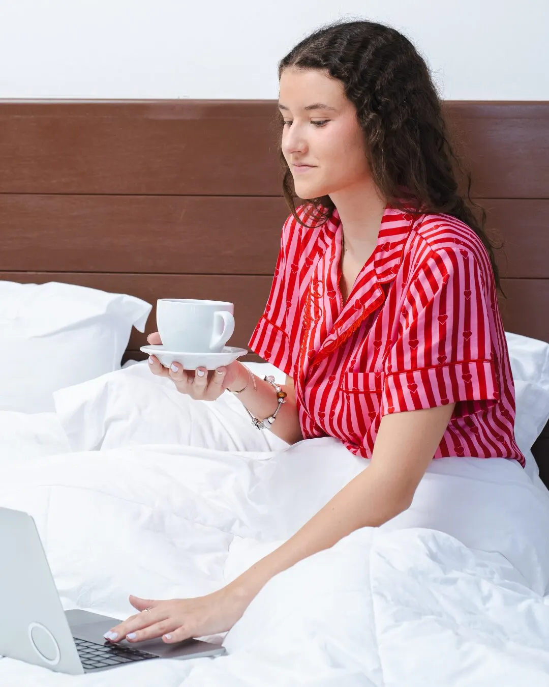 A young woman in red striped pajamas on a bed with a cup of coffee and a laptop.