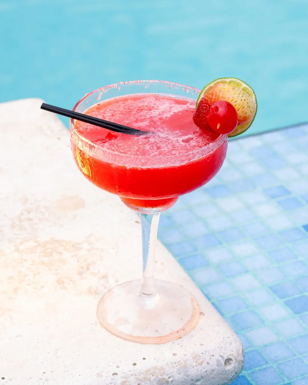 A red cocktail with a cherry and a lime wedge, on the edge of a pool.